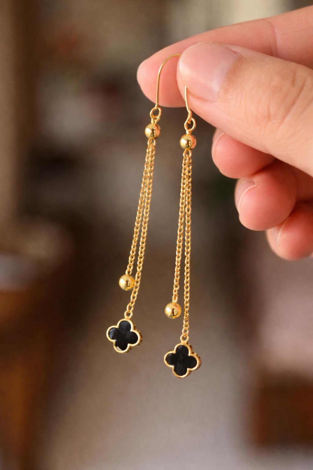 Gold earrings with black clover charms held by a hand against a blurred background