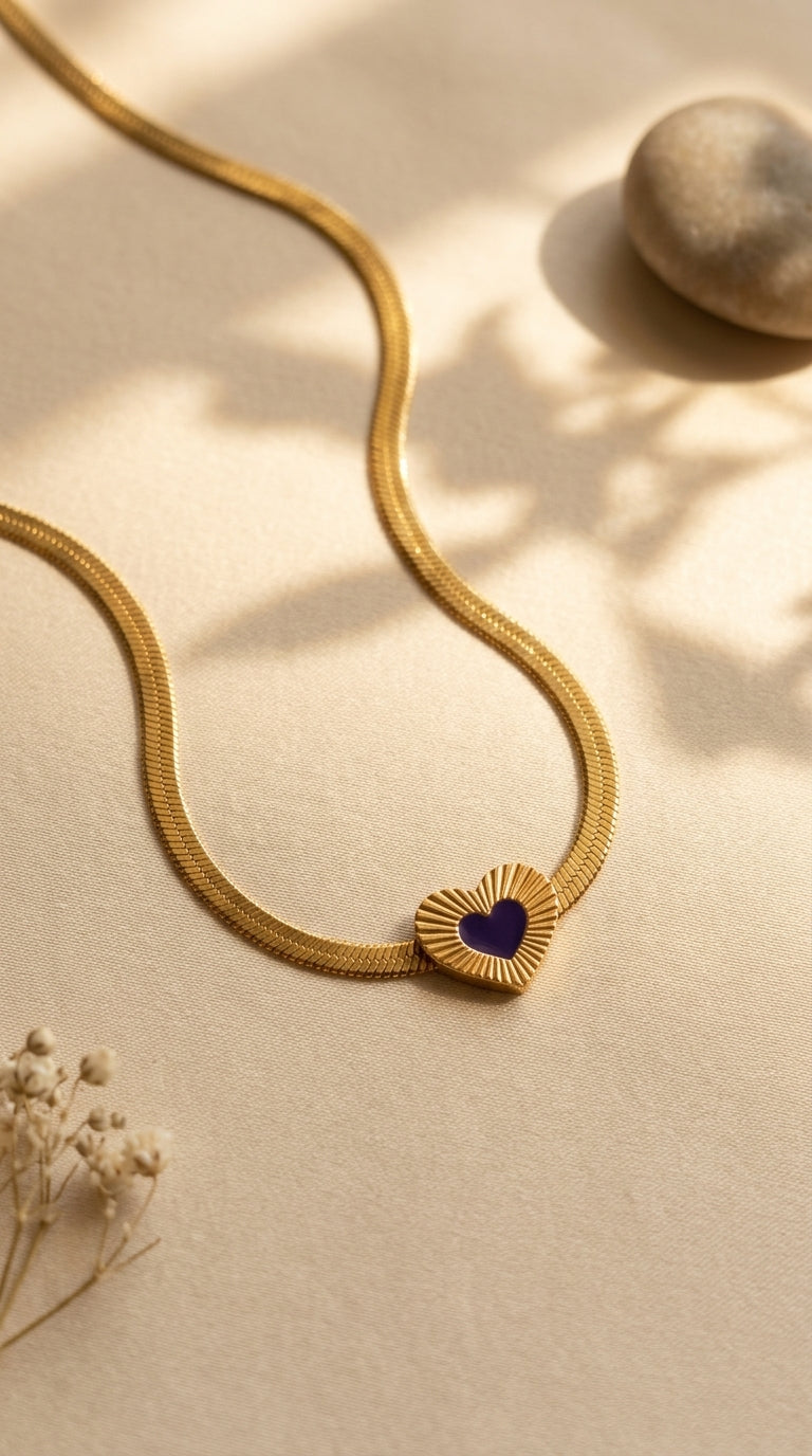 Gold necklace with a heart-shaped pendant on a beige surface with dried flowers and a rock.